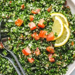 Tabbouleh salad in a white bowl with serving spoons and lemon slices