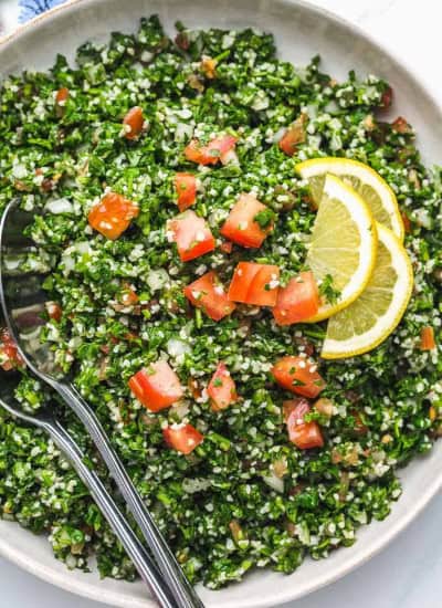 Tabbouleh salad in a white bowl with serving spoons and lemon slices