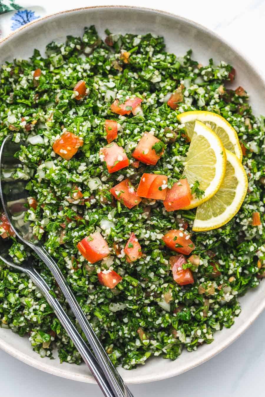 Tabbouleh salad in a white bowl with serving spoons and lemon slices
