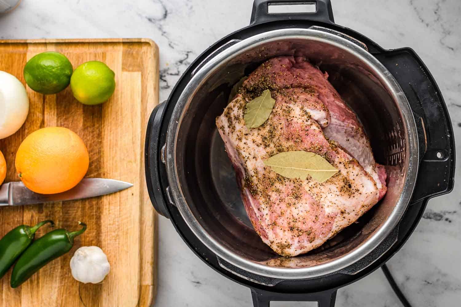 Pork shoulder in the instant pot, and a cutting board with limes, oranges, jalapeños, garlic.