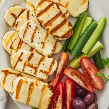 Grilled halloumi on a plate with sliced cucumber, tomatoes, and olives