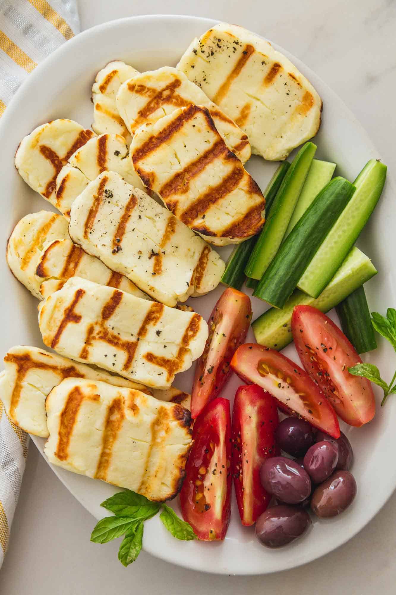 Grilled halloumi on a plate with sliced cucumber, tomatoes, and olives