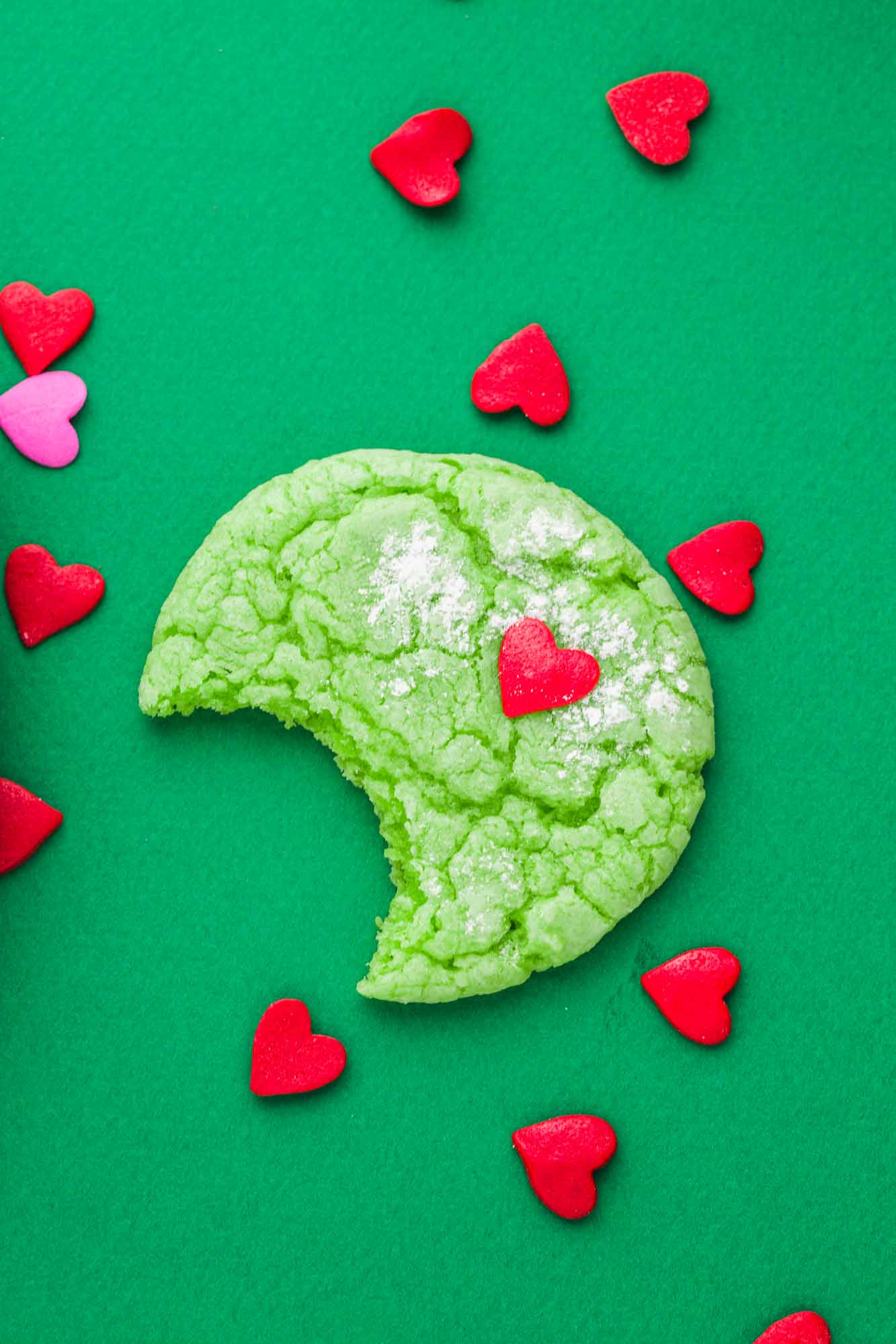 One green grinch cookie showing a bite shot, and heart sprinkles surrounding it. Placed on a dark green background.