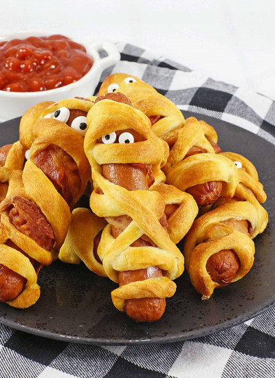 Halloween mummy hotdogs on a black plate with a bowl of ketchup in the background