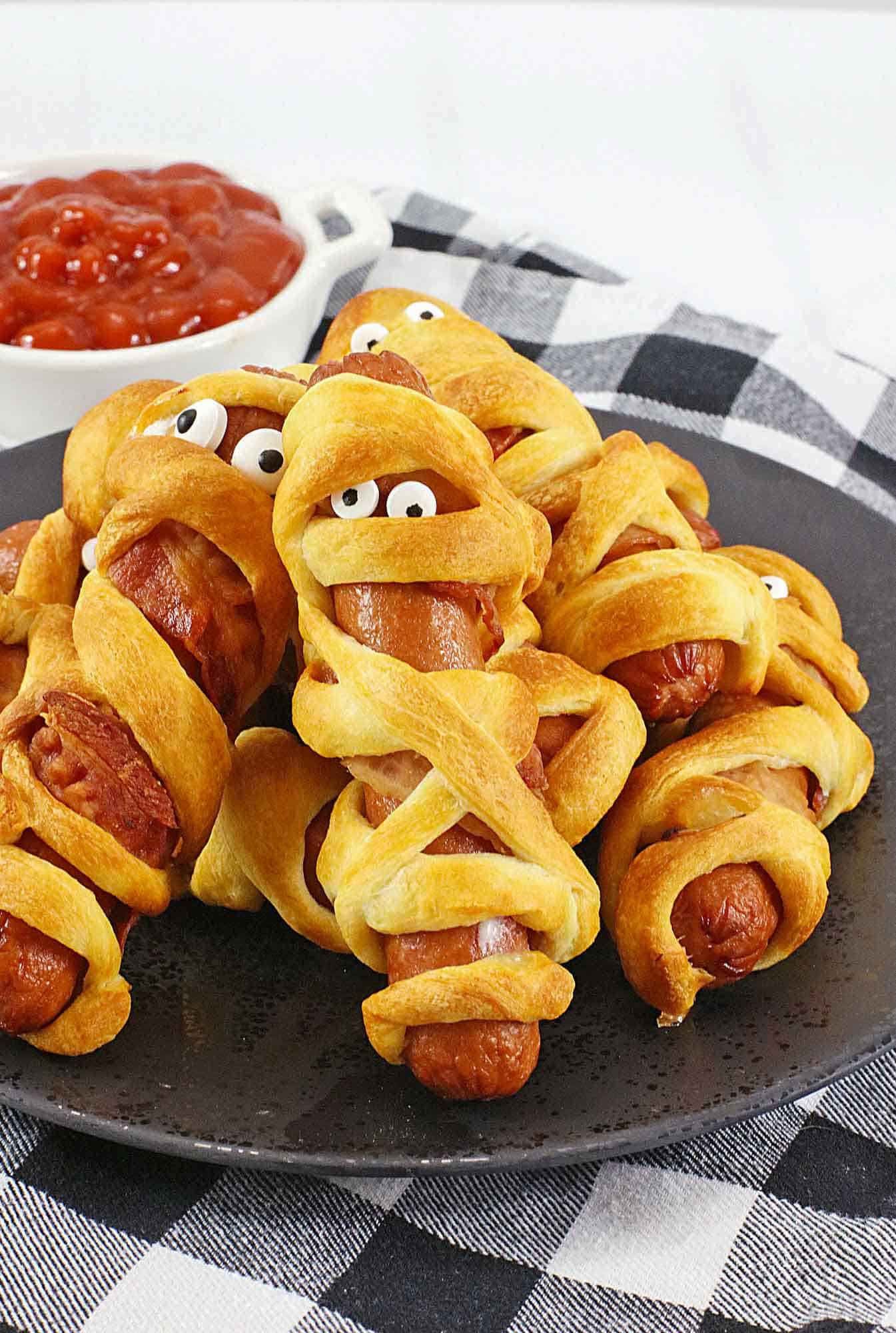 Halloween mummy hotdogs on a black plate with a bowl of ketchup in the background