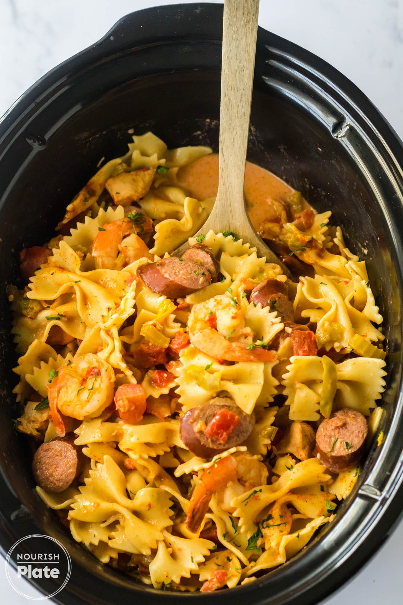 Overhead shot of jambalaya pasta in a black slow cooker with a wooden spoon