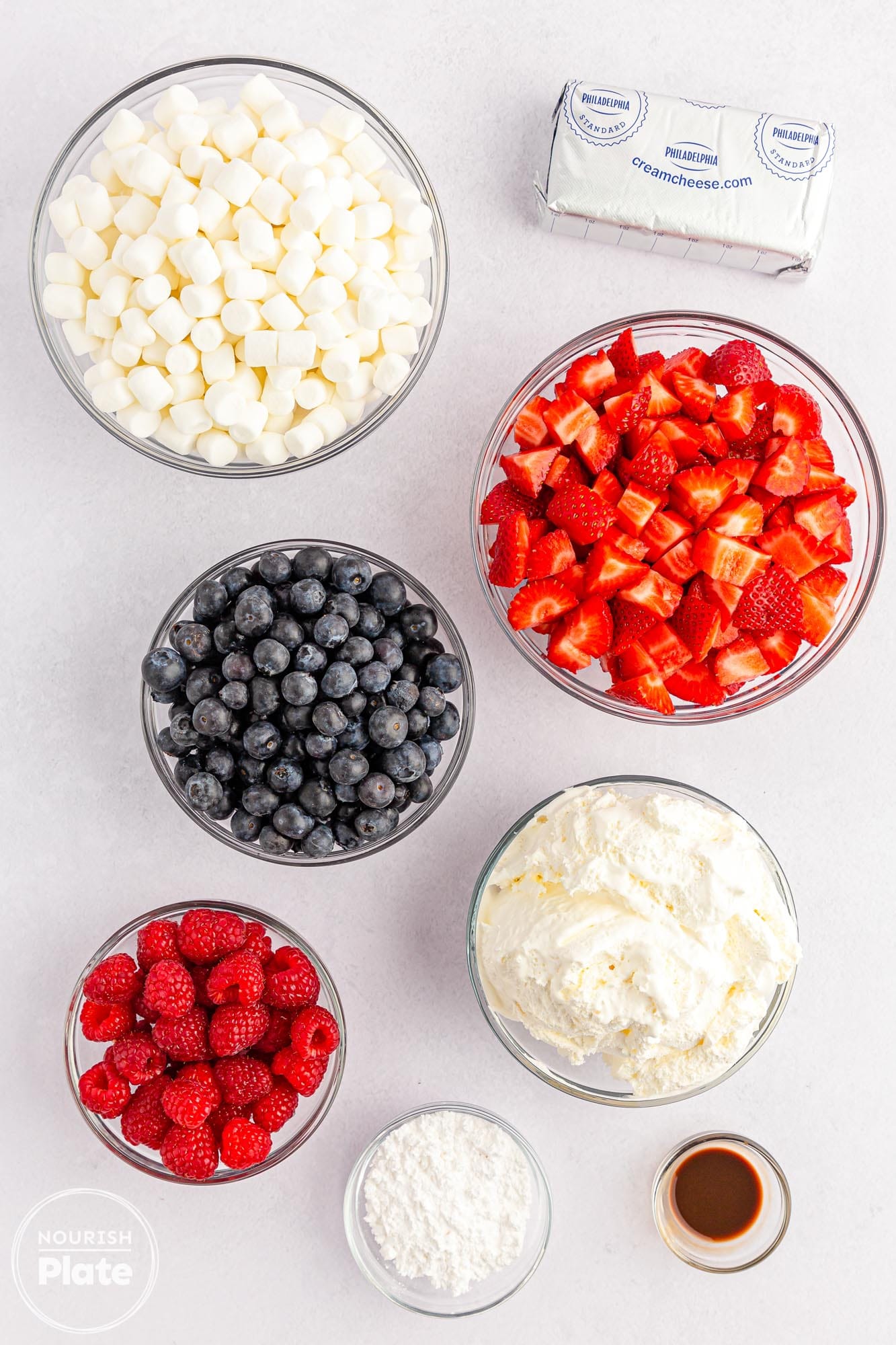 Overhead shot of cheesecake salad ingredients including whipped topping, cream cheese, fresh berries, mini marshmallows, and vanilla.