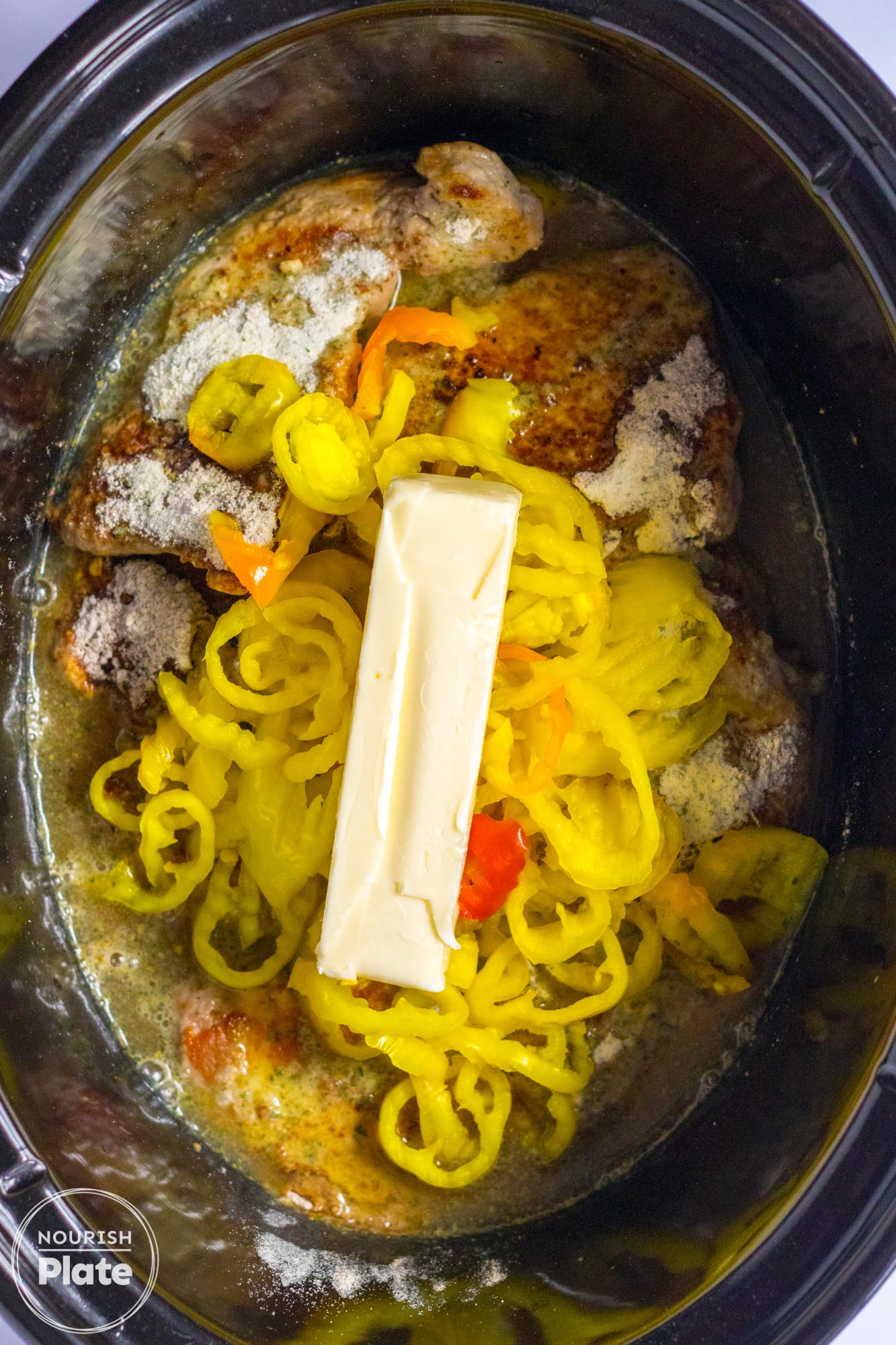Boneless pork chops in a black slow cooker, with peppers and a stick of butter before cooking
