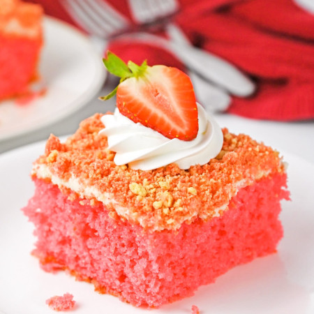 A slice of strawberry crunch cake with whipped cream on top and half a strawberry, plated on a white plate and 2 forks in the background with a red tea towel.