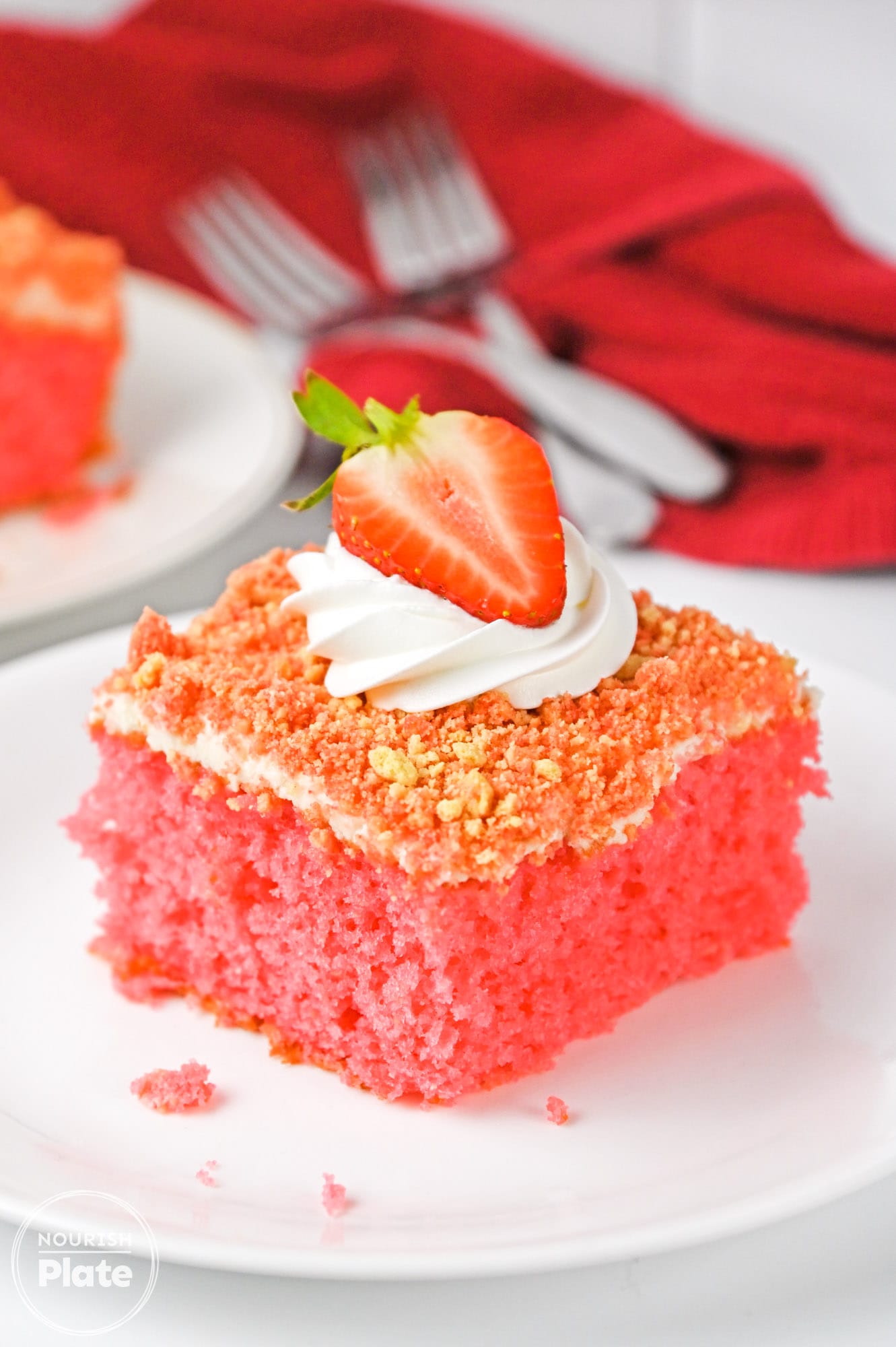 A slice of strawberry crunch cake with whipped cream on top and half a strawberry, plated on a white plate and 2 forks in the background with a red tea towel.