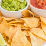 Crispy tortilla chips on a white platter, with 2 dips in the background (guacamole, and salsa)