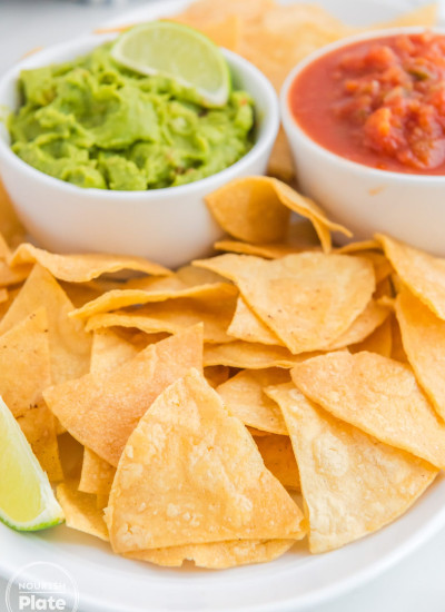 Crispy tortilla chips on a white platter, with 2 dips in the background (guacamole, and salsa)