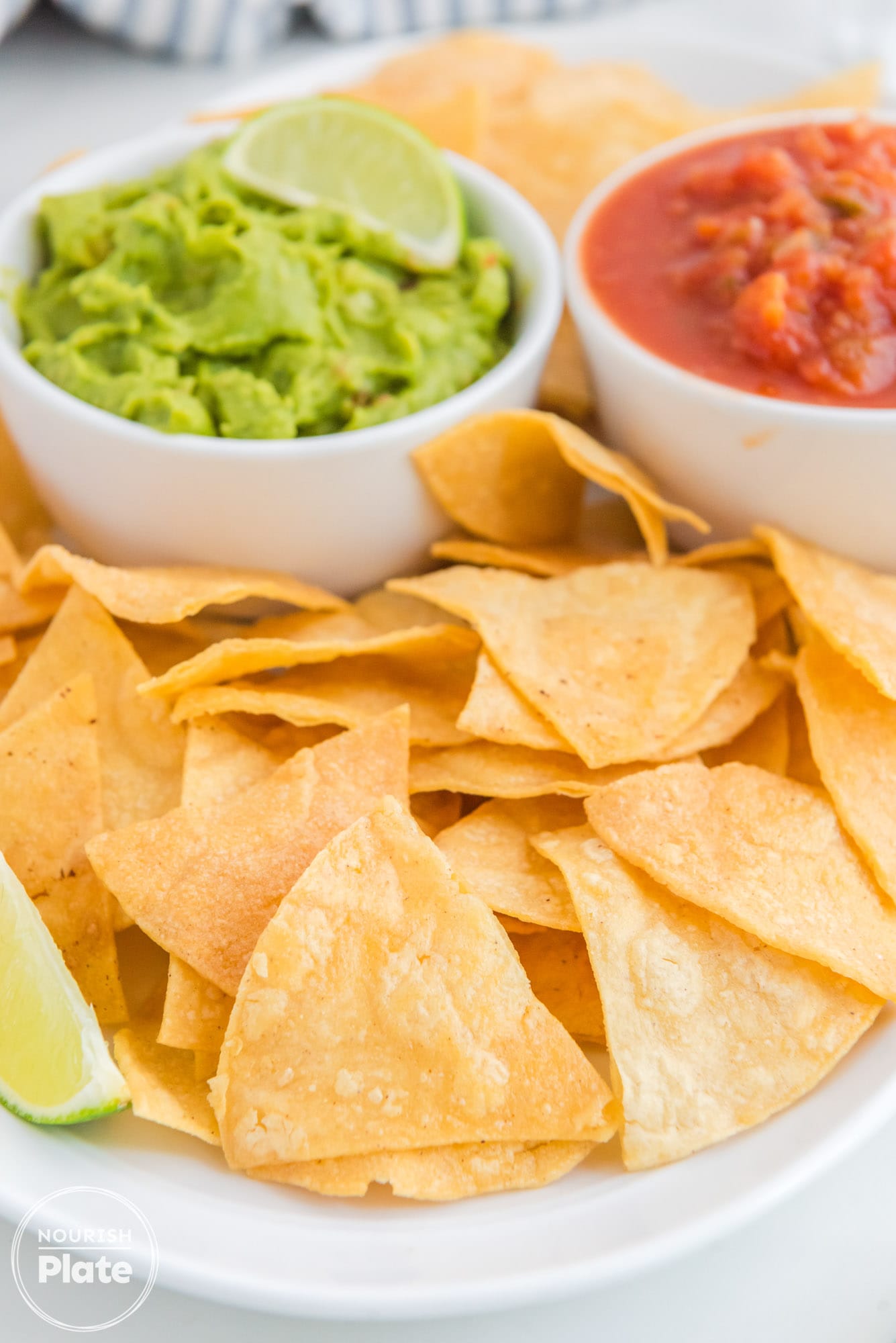 Crispy tortilla chips on a white platter, with 2 dips in the background (guacamole, and salsa)