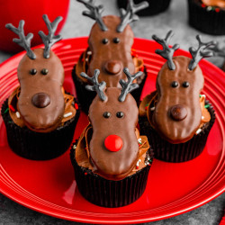 Chocolate cupcakes decorated with reindeer-shaped toppers made from chocolate-covered cookies, candy eyes, red and brown candies for noses, and antlers on a red plate. A red mug and additional cupcakes are in the background.