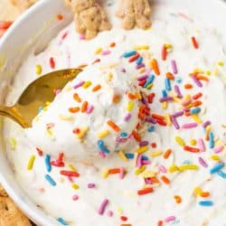 A close up of a white bowl filled with creamy white dip covered in colorful rainbow sprinkles, a gold spoon scooping the dip, and small teddy shaped graham crackers and sliced strawberries arranged around the bowl.