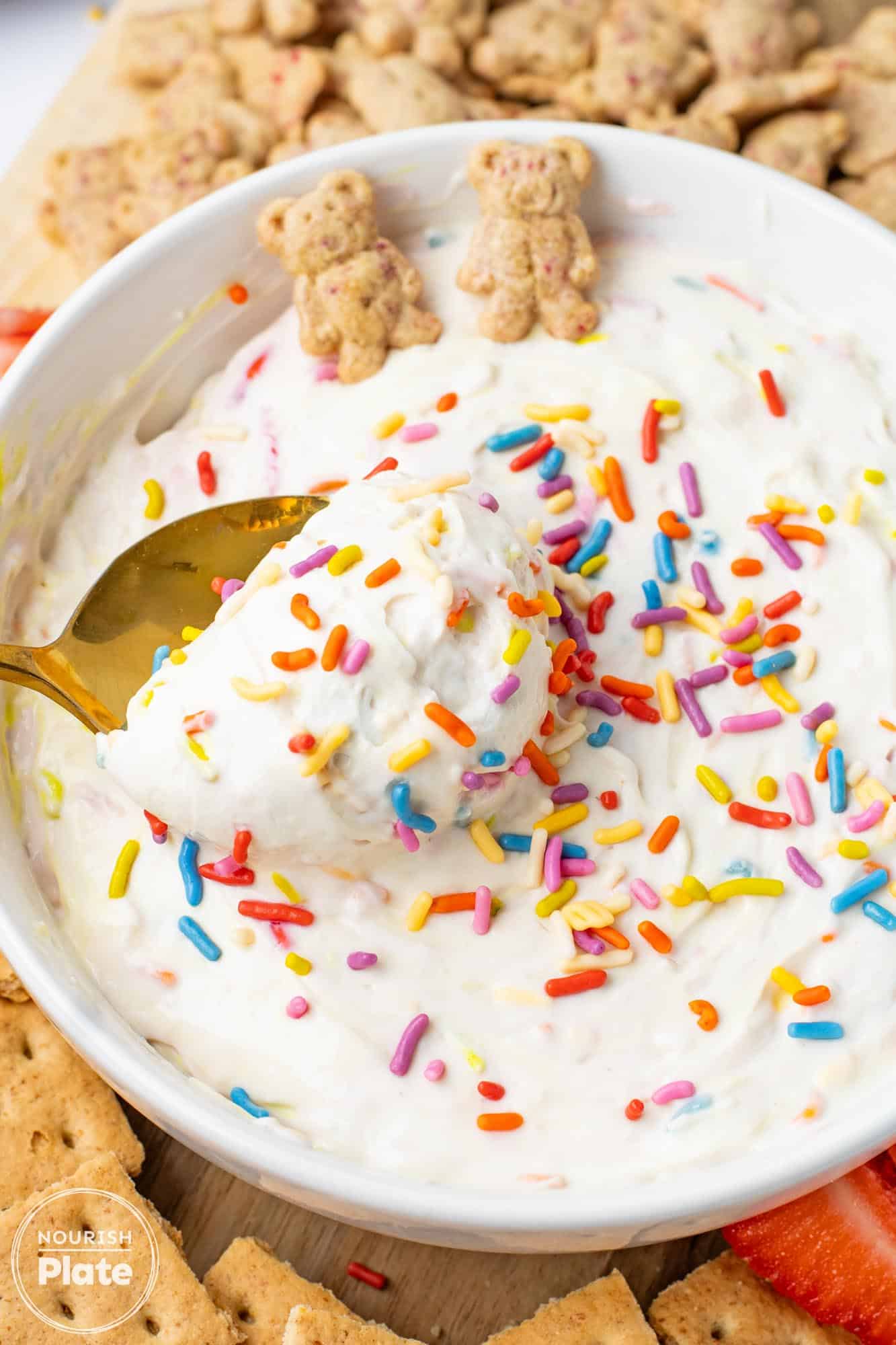 A close up of a white bowl filled with creamy white dip covered in colorful rainbow sprinkles, a gold spoon scooping the dip, and small teddy shaped graham crackers and sliced strawberries arranged around the bowl.