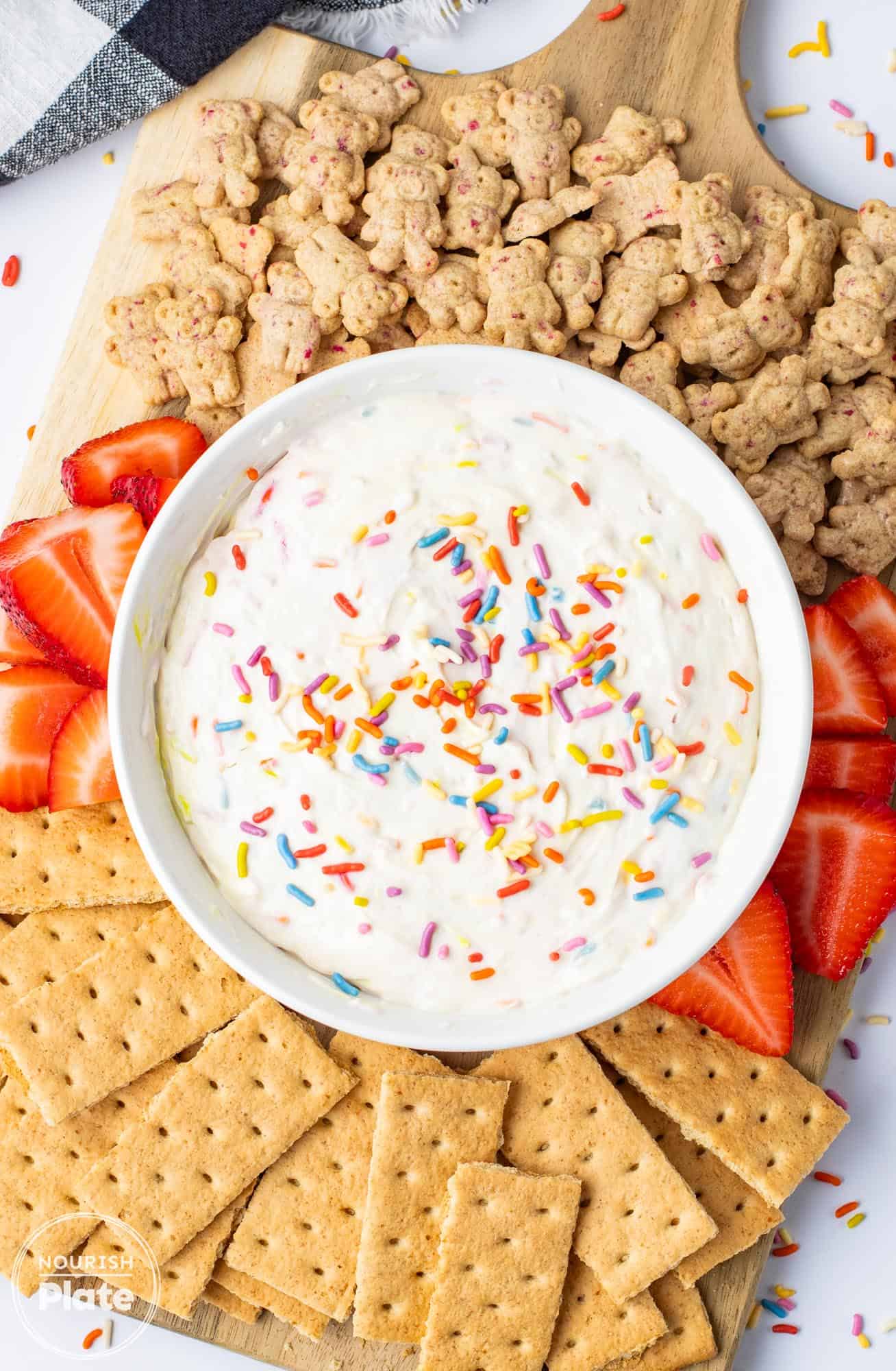 Overhead shot of dunkaroo dip in a white bowl, with graham teddies, fresh strawberries, and graham crackers on the side.