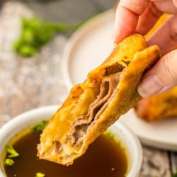 Close-up of a hand holding a golden-brown French Dip egg roll half, showing layers of roast beef, melted provolone, and onions as it’s dipped into a ramekin of dark au jus garnished with parsley.