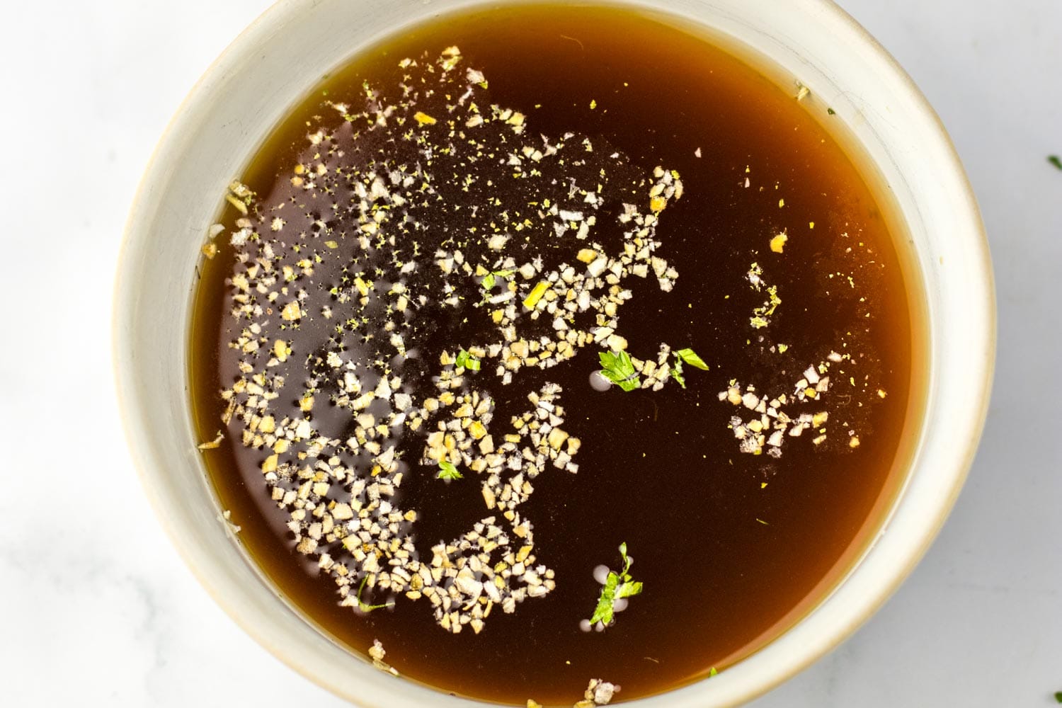Overhead view of dark beef broth in a small bowl, topped with dried minced onion and bits of parsley.