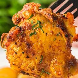 A close-up of a crispy air-fried chicken thigh held by a fork, with golden skin flecked with herbs and visible char marks.