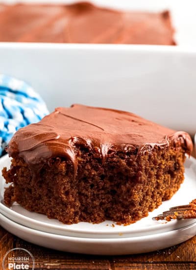 A slice of moist chocolate depression cake topped with a thick layer of chocolate frosting, served on a white plate with a fork. The background shows the rest of the frosted cake in a white baking dish.