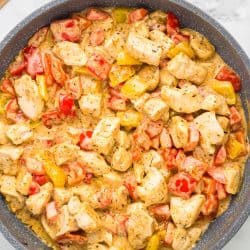 Top view of a large skillet filled with creamy Cajun chicken and sautéed bell peppers, next to a bowl of plain pasta and a wooden board with fresh parsley.
