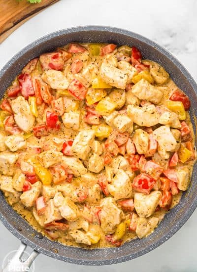 Top view of a large skillet filled with creamy Cajun chicken and sautéed bell peppers, next to a bowl of plain pasta and a wooden board with fresh parsley.