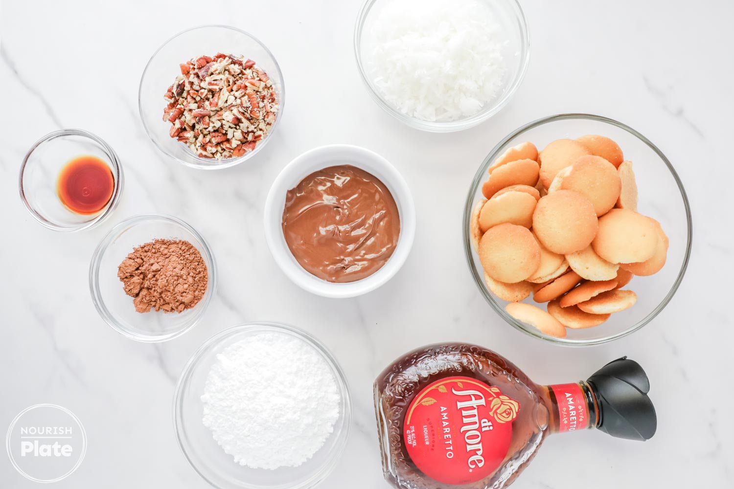 Overhead view of Amaretto ball ingredients arranged in glass bowls on a white surface, including vanilla wafers, chopped pecans, shredded coconut, powdered sugar, cocoa powder, Nutella, pure vanilla extract, and a bottle of Amaretto liqueur.