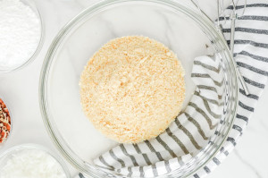 Glass bowl filled with finely crushed vanilla wafers, sitting on a striped towel with other ingredients visible around the edges.