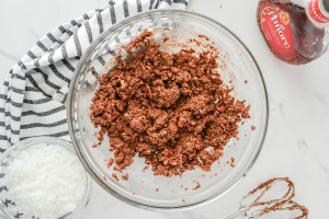Bowl of combined chocolate Amaretto ball mixture with a crumbly texture, placed on a striped towel with a bottle of Amaretto nearby.
