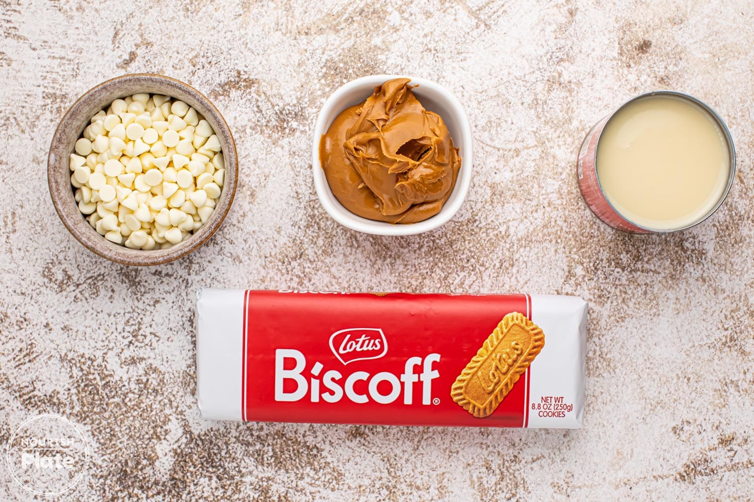 Overhead photo showing ingredients for cookie butter fudge: a bowl of white chocolate chips, a small dish of Biscoff cookie butter, a can of sweetened condensed milk, and a package of Lotus Biscoff cookies on a textured surface.