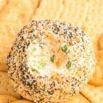 A round Everything Bagel Cheese Ball coated with sesame seeds, poppy seeds, garlic, and onion seasoning, with a creamy cheese and scallion filling visible in the center, surrounded by buttery Ritz and Club crackers for serving.