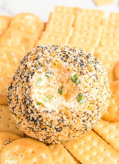 A round Everything Bagel Cheese Ball coated with sesame seeds, poppy seeds, garlic, and onion seasoning, with a creamy cheese and scallion filling visible in the center, surrounded by buttery Ritz and Club crackers for serving.