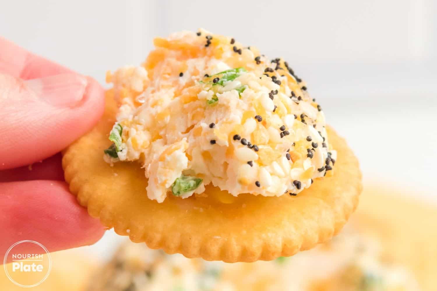 A close-up of a Ritz cracker topped with a scoop of creamy Everything Bagel Cheese Ball, showing bits of cheddar, scallions, and a coating of sesame seeds, poppy seeds, and garlic flakes.