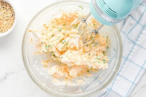 A mixing bowl filled with a creamy cheese mixture of cream cheese, shredded cheddar, and chopped scallions being blended with an electric hand mixer.