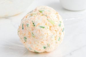 A smooth cheese ball coated with Everything Bagel seasoning resting on a plate covered with extra seasoning.