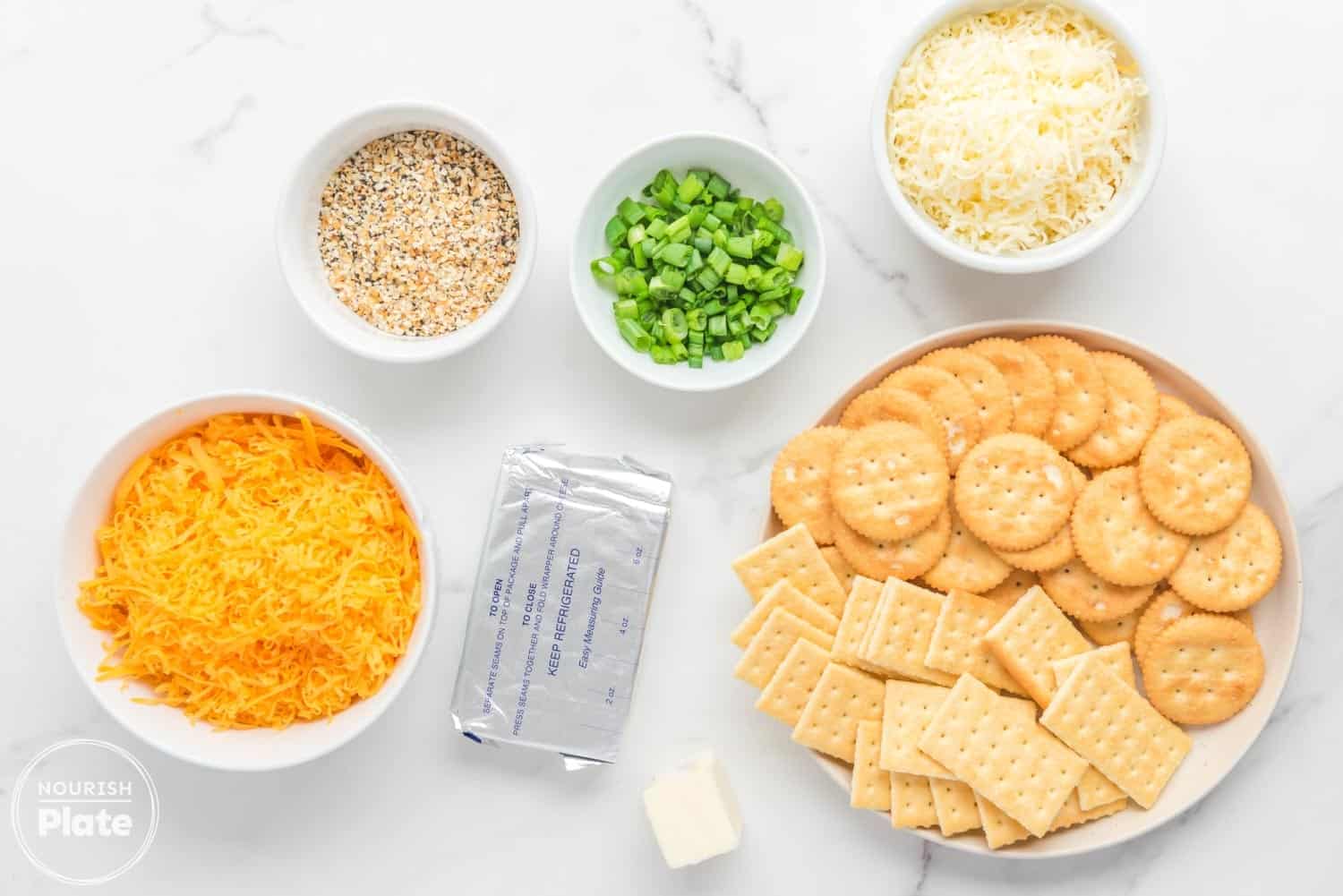 Overhead view of ingredients for Everything Bagel Cheese Ball, including shredded cheddar and colby jack cheeses, chopped scallions, Everything Bagel seasoning, a block of cream cheese, a small piece of butter, and a plate of Ritz and Club crackers on a white marble surface.