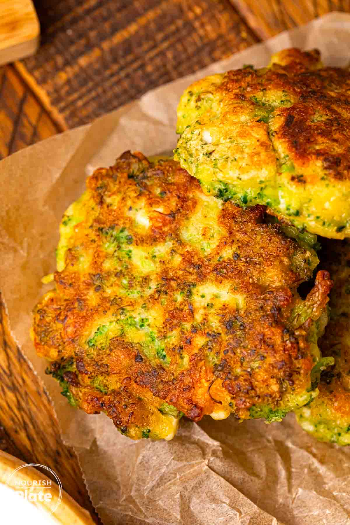 Close up of crispy pan fried broccoli fritters stacked on parchment paper, showing a golden brown crust and green broccoli pieces inside.