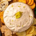 Mississippi Sin Dip in a glass bowl topped with chopped pepperoncini, surrounded by crackers, chips, and sliced bread on a wooden board