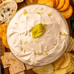 Mississippi Sin Dip in a glass bowl topped with chopped pepperoncini, surrounded by crackers, chips, and sliced bread on a wooden board