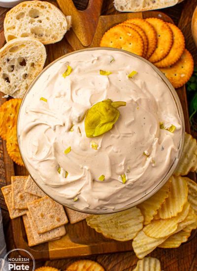 Mississippi Sin Dip in a glass bowl topped with chopped pepperoncini, surrounded by crackers, chips, and sliced bread on a wooden board