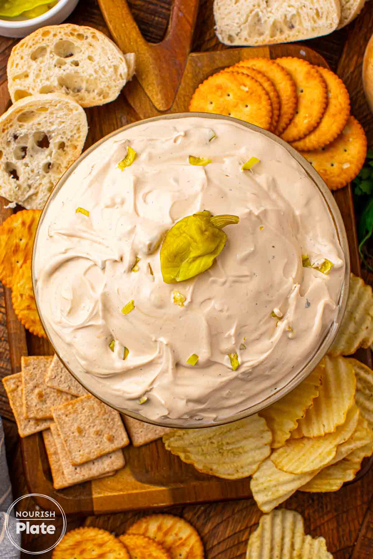 Mississippi Sin Dip in a glass bowl topped with chopped pepperoncini, surrounded by crackers, chips, and sliced bread on a wooden board