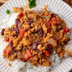 Cajun chicken with kidney beans, diced red bell pepper, and onion in a saucy Cajun style mixture spooned over white rice on a white rimmed plate, with fresh parsley in the background.