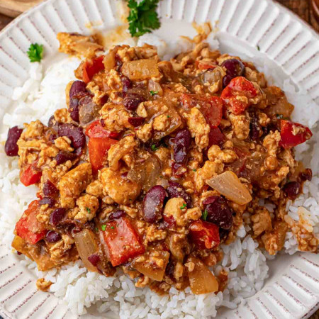 Cajun chicken with kidney beans, diced red bell pepper, and onion in a saucy Cajun style mixture spooned over white rice on a white rimmed plate, with fresh parsley in the background.