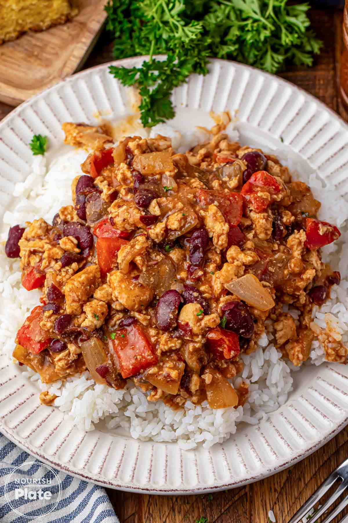 Cajun chicken with kidney beans, diced red bell pepper, and onion in a saucy Cajun style mixture spooned over white rice on a white rimmed plate, with fresh parsley in the background.