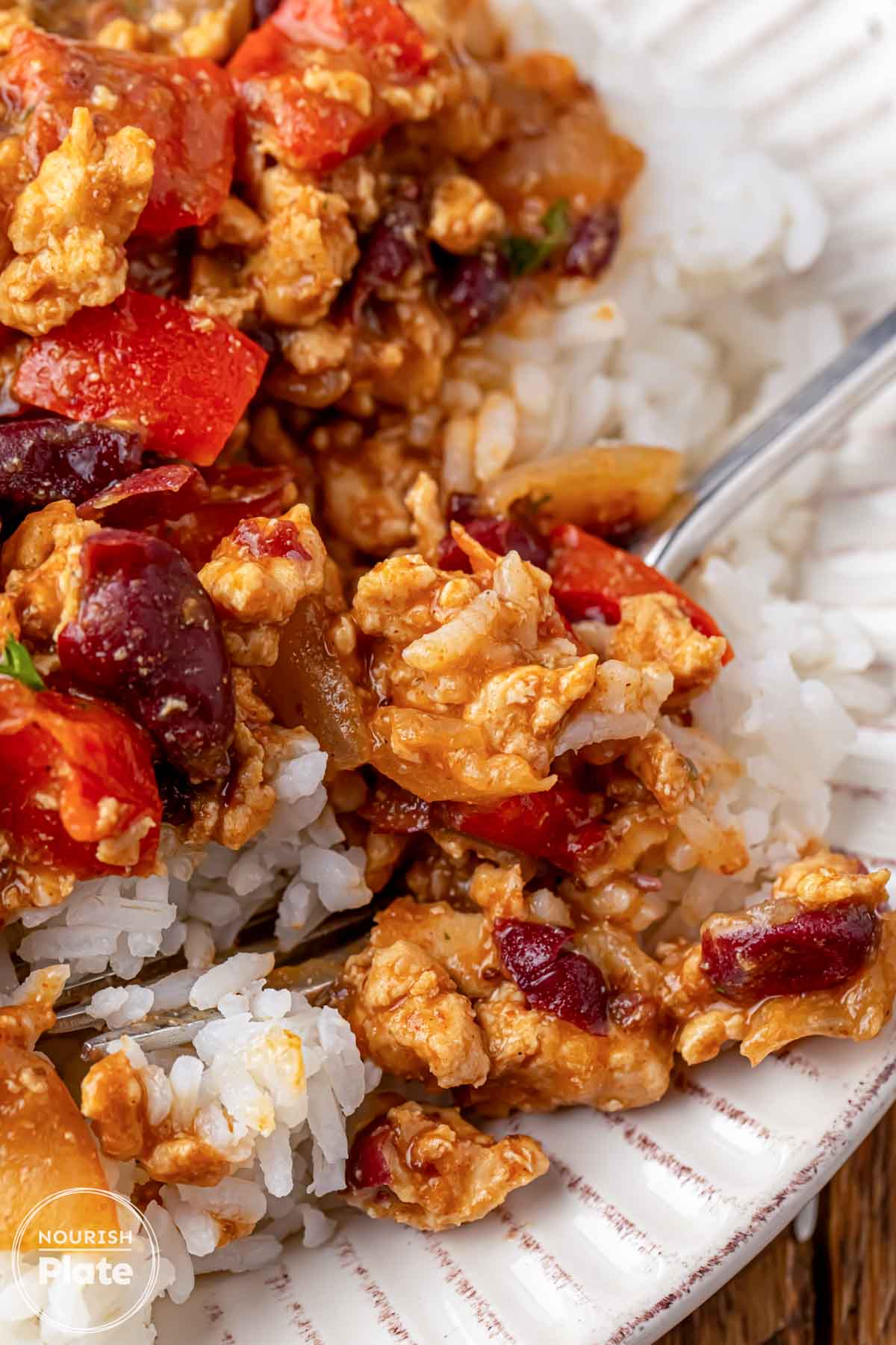 Close up of Cajun ground chicken and kidney beans with red bell pepper and onion over white rice, with a spoon scooping a bite from the plate.