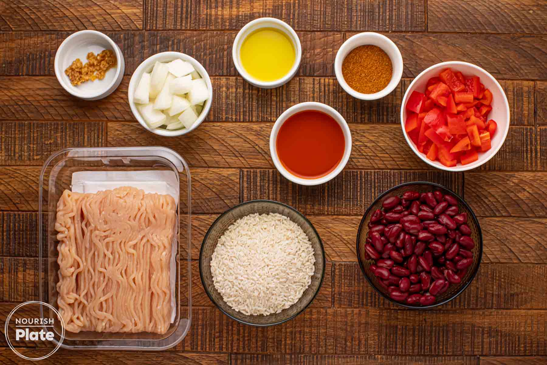 Overhead view of ingredients for Cajun chicken red beans and rice including ground chicken, kidney beans, white rice, diced onion, chopped red bell pepper, minced garlic, Cajun seasoning, olive oil, hot sauce, and broth on a wooden board.