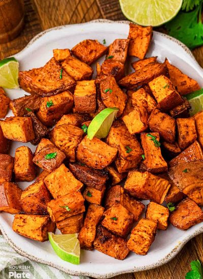 Roasted sweet potato cubes seasoned with chili and smoked paprika on a white plate, garnished with lime wedges and chopped cilantro.