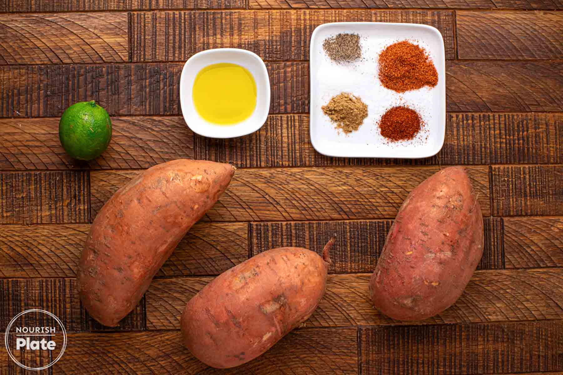Overhead view of sweet potatoes, a whole lime, a bowl of olive oil, and a plate of chili powder, smoked paprika, cumin, and black pepper on a wooden board.