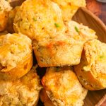 Close up of beer cheese bread muffins piled on a wooden plate, topped with melted cheddar and bits of green chile and chives.